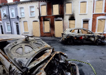 Charred, burned-out cars on a residential street with boarded-up windows in a row of terraced houses