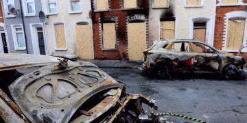 Charred, burned-out cars on a residential street with boarded-up windows in a row of terraced houses