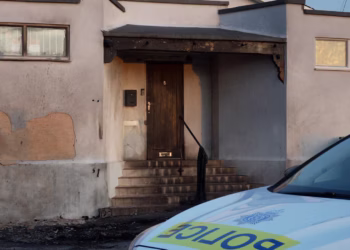Entrance to a burned building with charred wooden beams, steps, and a police car in the foreground